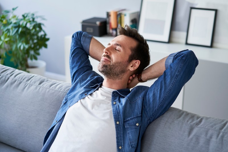 Un hombre con los ojos vendados descansa en un sofá gris, con las manos detrás del cuello.