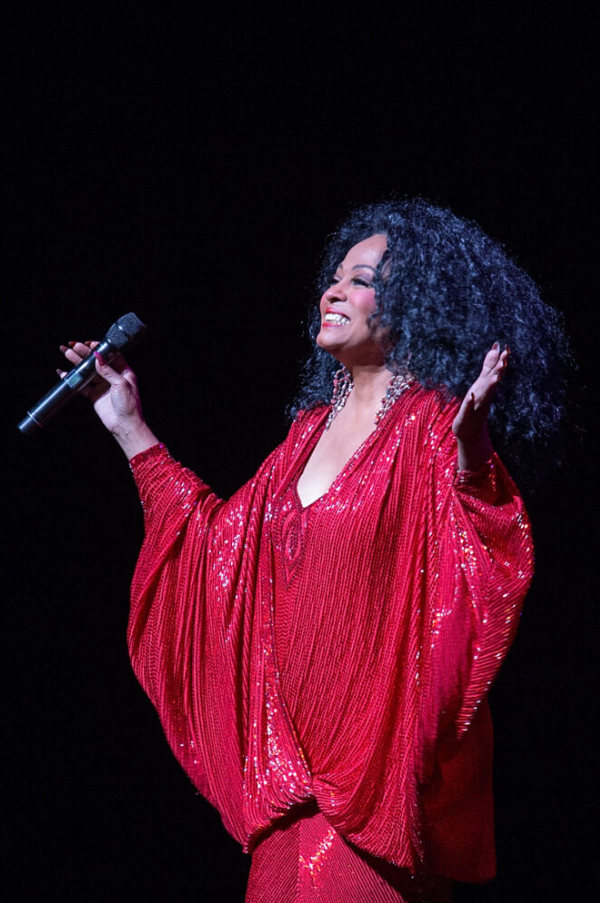 Diana Ross con un vestido rojo brillante que sostiene un micrófono, sonríe y sostiene sus brazos