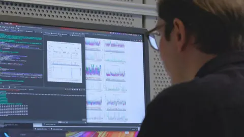 Jeff Dowling / Embl-Ebi, la parte posterior de la cabeza y los hombros del hombre de cabello oscuro mientras mira la pantalla de una computadora. Se puede ver la imagen de un código de computadora y gráficos multicolores, aunque no se conoce su significado. 