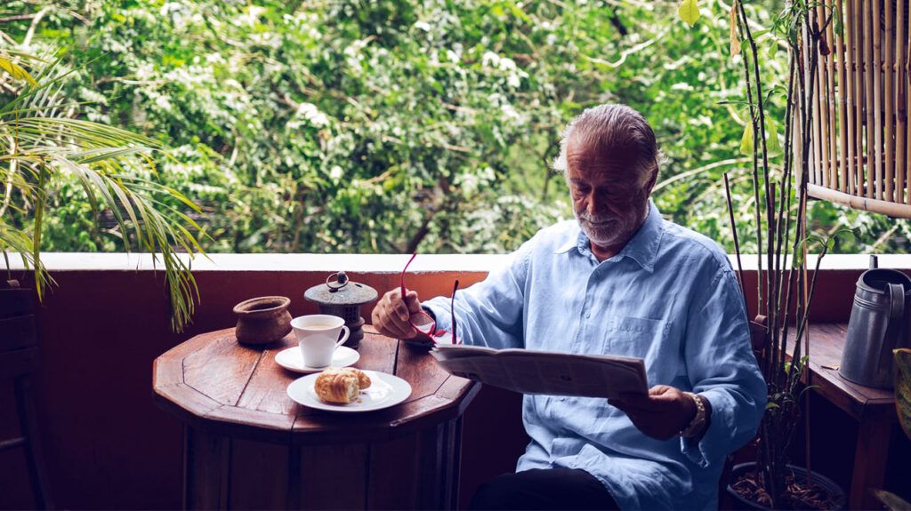 Un viejo café adulto canta un periódico con las gafas que están en manos de una mesa cerca de una mesa cerca de una mesa cerca de una mesa y una mesa al lado de una ventana