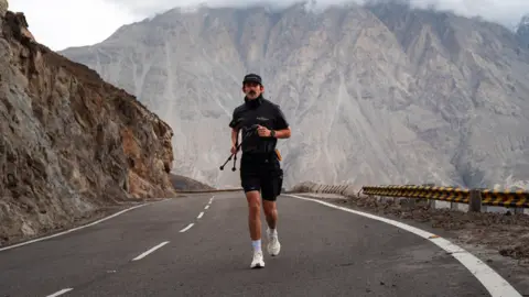 Foto de Jack Paisley trabajando en una carretera en la lado de la montaña. Las grandes montañas se sientan hacia atrás mientras ejecutan todo el material negro hacia la cámara.