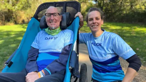 BBC David y Alice tomaron una foto en un parque. David tiene cabello gris corto, gafas negras nutritivas y una perilla. Se sienta en una silla de ruedas lisa. Alice cerró el cabello castaño claro y se dobla al lado de la silla.