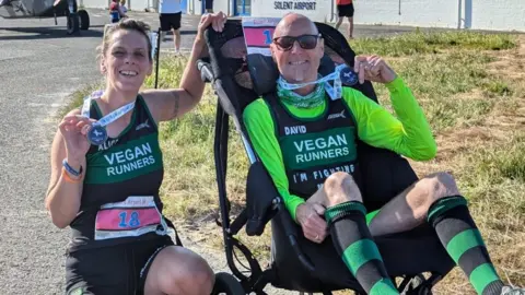 David y Alice sostienen medallas después de correr en el aeropuerto de Solent. Alice David está con una silla de ruedas. Ambos usan ropa y sonriendo en la cámara.