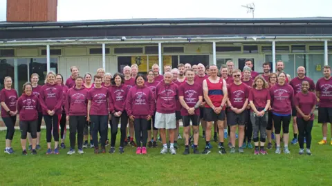 Un gran grupo de Saxon Running & Fitness Club se encuentra en un deporte de hierba fuera de un pabellón. Todos usan titulares de color púrpura oscuro con un logotipo de White Clubs en la primera línea. Sonríen en la cámara.
