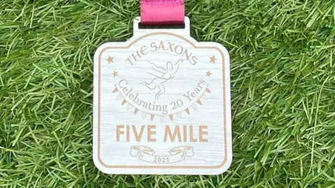 Sasons celebró en el Running & Fitness Club grabado y leído en una medalla: Sevenons, 20 años, cinco millas, 2025. 