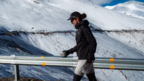 El golpe de Devin Paisley, que es la vista de Jack, que es un equipo que huye en un pase de montaña cubierto de nieve. Usa una taza de café en el guante, el sombrero y la mano derecha 