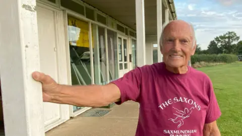 Sajones Running & Fitness Club Gordon Mercield, una mano en la apariencia de un pabellón deportivo está sonriendo en la cámara mientras una mano descansa una mano en el exterior de un pabellón deportivo. Tiene un poco de cabello gris y usa una camisa morada oscura con secciones con el logotipo del club. Su hierba a su derecha tiene un terreno deportivo.
