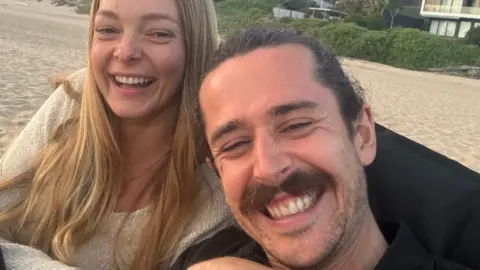 Selfie débil tomada por Jack y Bronwyn. Sonríen y se sientan en una playa