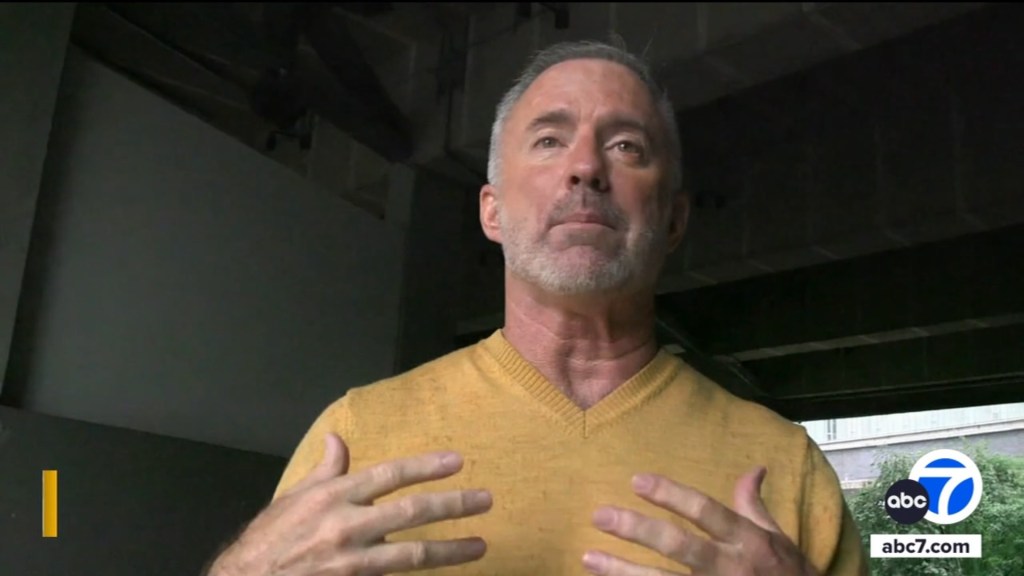 Un hombre de cabello gris y barba que viste un suéter amarillo y gesticula con las manos.