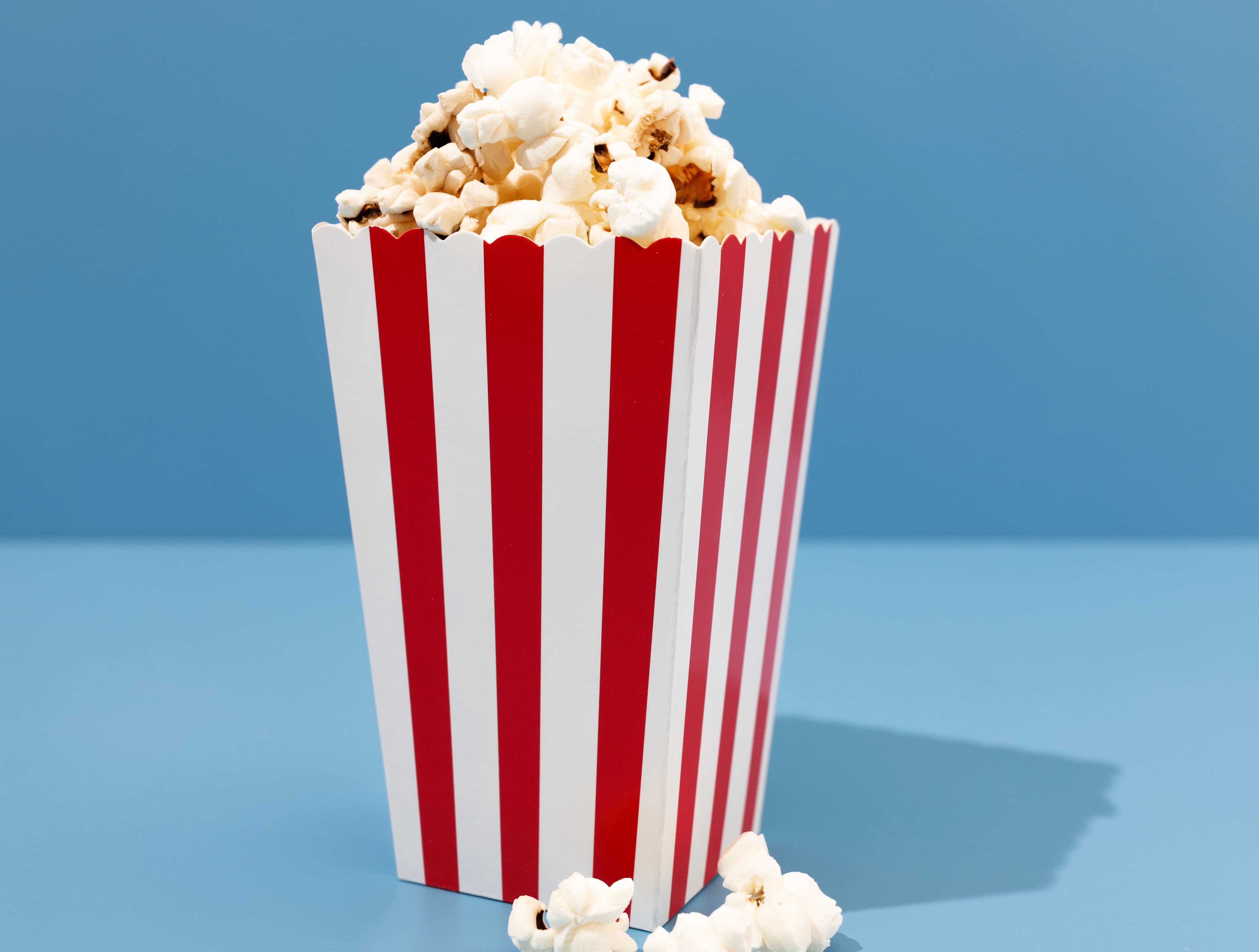 Un cubo de palomitas de maíz a rayas rojas y blancas lleno de palomitas de maíz, sobre un fondo azul.