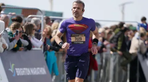 Toby Phillips Photography, un hombre con una camiseta deportiva morada y pantalones cortos azul marino. Lleva zapatillas deportivas de color azul brillante. Trabaja en una carrera con un gran número de espectadores al lado.