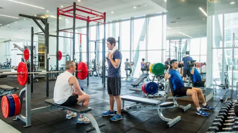 Getty Images La gente hace ejercicio en lo que parece ser un gimnasio comercial. Un hombre se sienta en un banco de pesas y habla con un hombre que está frente a él. En la esquina derecha, un hombre levanta pesas con ambas manos. Al fondo se pueden ver mujeres en bicicleta. 