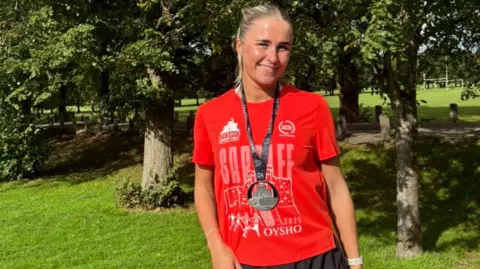 Leah Walsh Leah sonríe a la cámara. Se para frente a árboles y vegetación y viste una camiseta roja de finalista de la Media Maratón de Cardiff, una medalla negra y pantalones cortos negros para correr. Leah tiene el pelo rubio recogido en una coleta alta. 