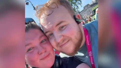 Un selfie en primer plano de Richard con su novia Sophie en el evento de carreras Richard Gallimore Race for Life. Sophie, a la izquierda, tiene cabello castaño y usa gafas de sol en la parte superior de la cabeza. Richard está de pie junto a Sophie, su mejilla tocando la de ella. Viste una camisa verde, una medalla rosa, auriculares y tiene el pelo rubio corto. 