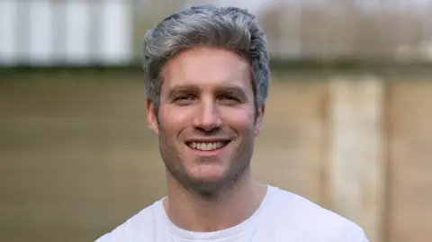 Michael James Primer plano de Dafydd sonriendo a la cámara. Está parado frente a una pared de ladrillos, pero el fondo está borroso. Viste una camisa blanca y tiene el pelo gris.
