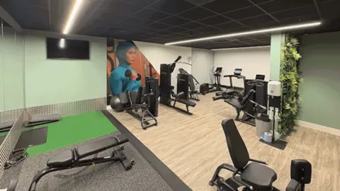 Un gimnasio exclusivo para mujeres tras la renovación del centro Everyone Active Recreation. La habitación tiene paredes de color verde claro y blanco con tiras de iluminación y un piso de madera clara, con una pequeña sección alfombrada de color gris. Hay varios tipos de equipos de gimnasio, incluidas elípticas y máquinas de remo. Hay algunas hojas artificiales en la pared, un televisor y grandes espejos, una estación de limpieza y un gran cuadro de una mujer levantando pesas.