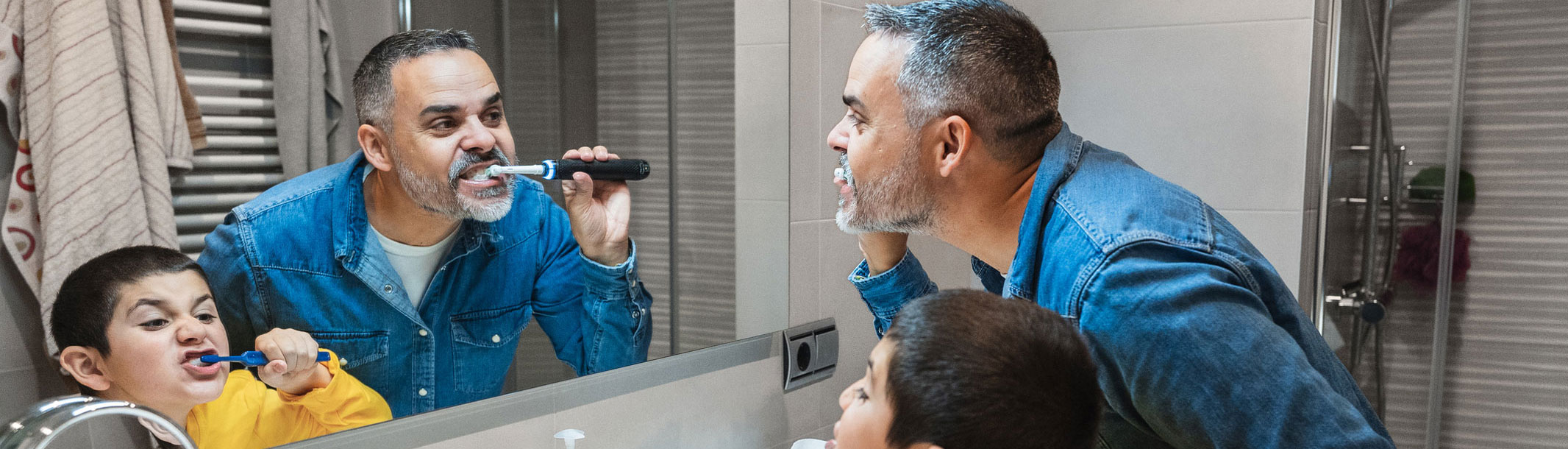 Foto de padre e hijo pequeño cepillándose los dientes con un cepillo eléctrico mientras están parados frente al espejo del baño