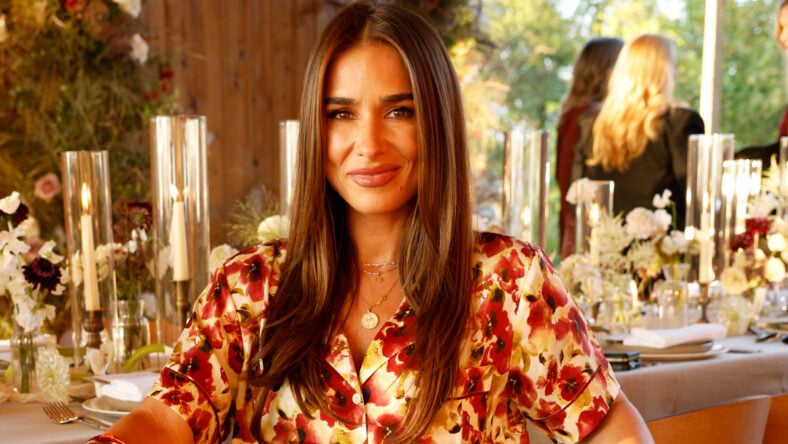 Jessie James Decker viste una blusa floral roja y amarilla y sonríe mientras está sentada en una mesa de comedor decorada con flores a la luz de las velas.